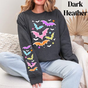 May include: A dark heather gray long-sleeve sweatshirt featuring a graphic print of colorful bats in shades of pink, orange, yellow, and light blue, interspersed with small stars. The design is repeated on the front torso and down the left sleeve.