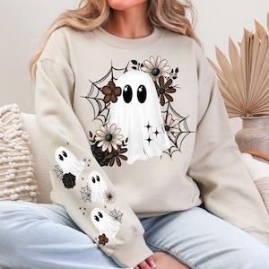 May include: Beige sweatshirt featuring a white ghost design with large black eyes, surrounded by brown and cream-colored flowers and spiderwebs. The sleeve has additional ghost and spiderweb graphics. A cozy, seasonal garment.