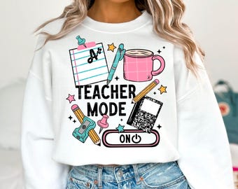 May include: White sweatshirt with a colorful graphic design. The design includes a pink mug, pencils, a notepad, and the words "Teacher Mode ON". The sweatshirt is a comfortable, casual style.