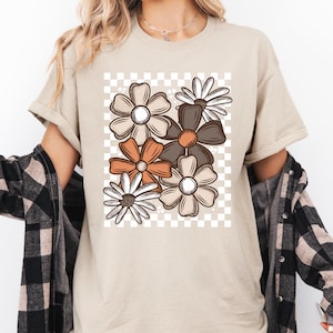 以下が含まれることがあります： ベージュのTシャツ。茶色、オレンジ、白の花柄がチェックの背景に配置されています。Tシャツは半袖で、ゆったりとしたフィット感です。柔らかい素材で作られています。