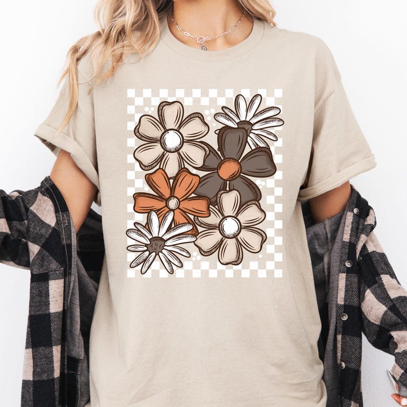 以下が含まれることがあります： ベージュのTシャツ。茶色、オレンジ、白の花柄がチェックの背景に配置されています。Tシャツは半袖で、ゆったりとしたフィット感です。柔らかい素材で作られています。