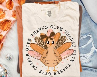 Könnte beinhalten: Ein cremefarbenes T-Shirt mit einer zentralen Grafik eines Cartoon-Truthahns mit brauner Schleife, umgeben von dem kreisförmig wiederholten Text "GIVE THANKS". Der Truthahn hat gemusterte Federn in Braun- und Rosatönen.