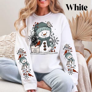 K&ouml;nnte beinhalten: Wei&szlig;es Sweatshirt mit Schneemann-Design, gr&uuml;ner M&uuml;tze, Schal und Kn&ouml;pfen. Die &Auml;rmel zeigen passende Schneemann- und Vogelgrafiken. Das Wort "White" ist oben rechts aufgedruckt. Ein festliches Winter-Bekleidungsst&uuml;ck.