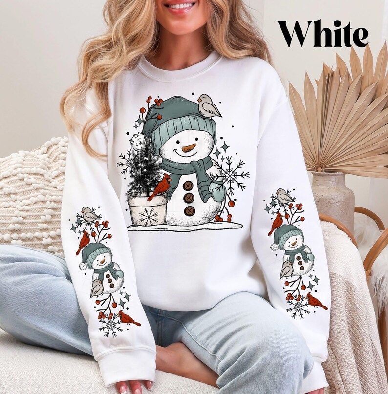 K&ouml;nnte beinhalten: Wei&szlig;es Sweatshirt mit Schneemann-Design, gr&uuml;ner M&uuml;tze, Schal und Kn&ouml;pfen. Die &Auml;rmel zeigen passende Schneemann- und Vogelgrafiken. Das Wort "White" ist oben rechts aufgedruckt. Ein festliches Winter-Bekleidungsst&uuml;ck.