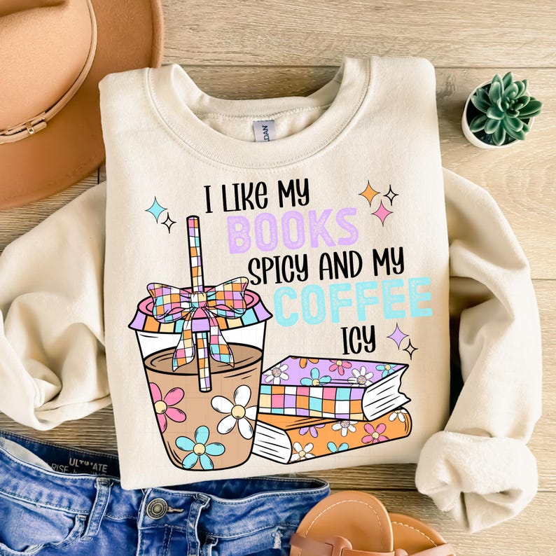 May include: A beige sweatshirt with the text "I like my books spicy and my coffee icy" printed on it. The text is surrounded by colorful stars and a cartoon illustration of a coffee cup with a straw and a bow, and a stack of books.