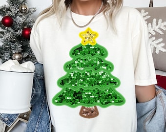 May include: Cream-colored t-shirt featuring a green sequined Christmas tree design with a yellow star on top. The tree has a brown trunk. The shirt is a festive holiday apparel item.