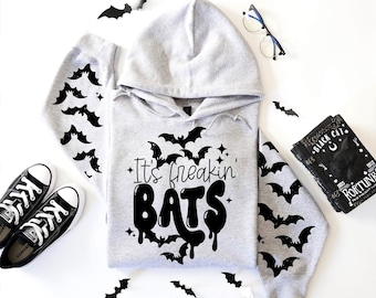 Könnte beinhalten: Ein hellgrauer Kapuzenpullover mit einem schwarzen Grafikdruck von fliegenden Fledermäusen und dem Text "It's freakin' BATS" auf der Vorderseite. Die Ärmel sind mit einem sich wiederholenden schwarzen Fledermausmuster verziert.