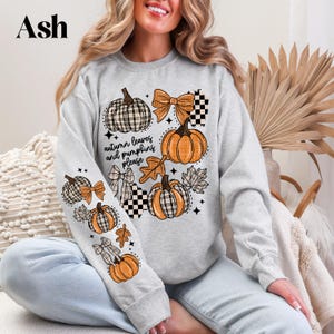 May include: A light gray sweatshirt with an autumn-themed graphic. The design features pumpkins, bows, and leaves in orange, black, and white. The text "autumn leaves and pumpkins please" is also included. The word "Ash" is in the top left corner.