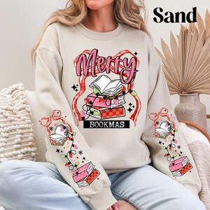 May include: A sand-colored sweatshirt with a festive "Merry BOOKMAS" design. The graphic features a stack of books, string lights, and a decorative ribbon. The sleeves also have book and light designs.