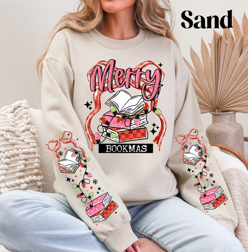 May include: A sand-colored sweatshirt with a festive "Merry BOOKMAS" design. The graphic features a stack of books, string lights, and a decorative ribbon. The sleeves also have book and light designs.
