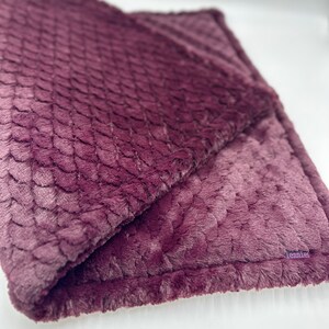 Op de afbeelding: Een zachte, pluche deken in een diepe paarse tint met een getextureerd, gestikt patroon. De deken is dubbelgevouwen en heeft een klein label met de tekst "Jennies".