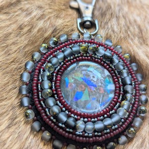 May include: A circular beaded keychain featuring a central cabochon with iridescent blue and green shards. The piece is surrounded by concentric rings of deep red and matte grey beads, finished with a silver-toned metal lobster clasp attachment.