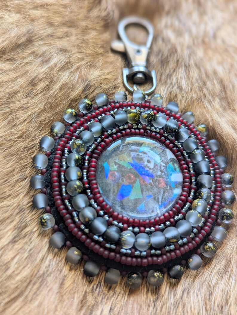 May include: A circular beaded keychain featuring a central cabochon with iridescent blue and green shards. The piece is surrounded by concentric rings of deep red and matte grey beads, finished with a silver-toned metal lobster clasp attachment.