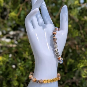May include: Two beaded bracelets displayed on a white ceramic hand. One bracelet features gold-colored beads and brown and tan patterned beads. The other bracelet has silver-colored beads and brown and tan patterned beads.
