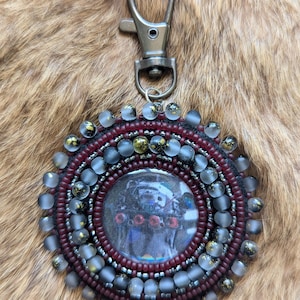 May include: A circular keychain featuring a central image of a dark figure with red accents, surrounded by concentric rings of deep maroon and translucent frosted beads with gold flecks, finished with a silver-toned metal lobster clasp.