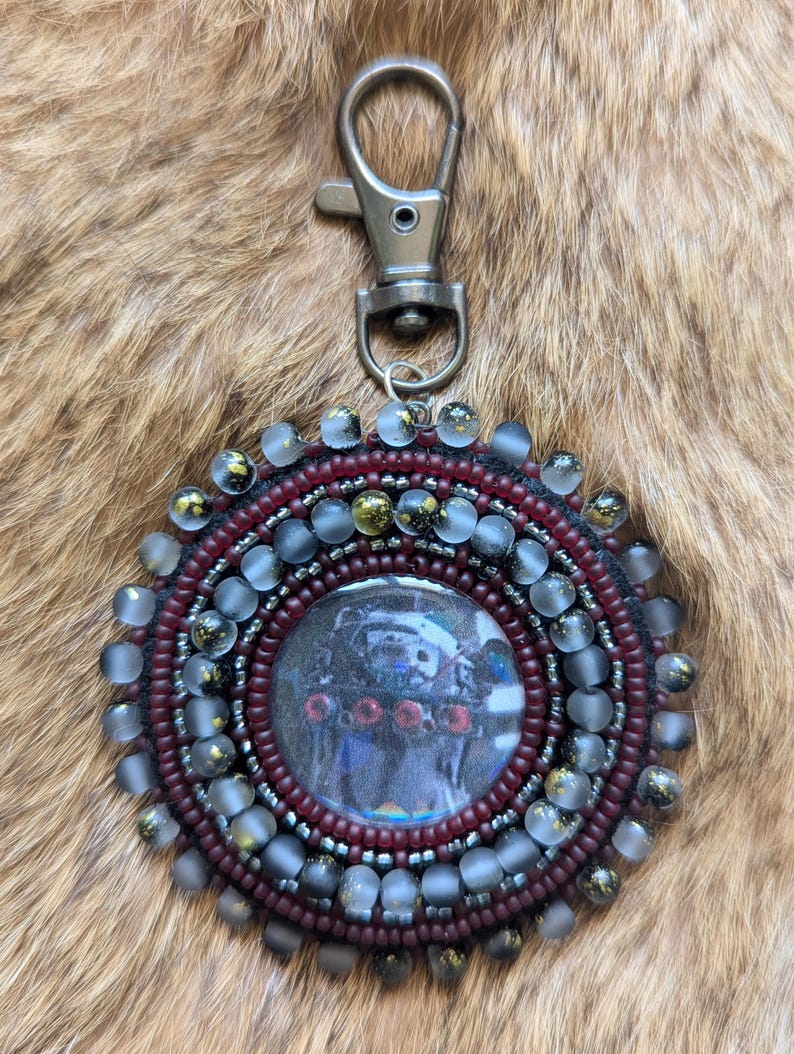 May include: A circular keychain featuring a central image of a dark figure with red accents, surrounded by concentric rings of deep maroon and translucent frosted beads with gold flecks, finished with a silver-toned metal lobster clasp.
