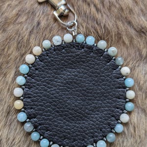 Op de afbeelding: Een ronde zwarte leren sleutelhanger met een zilveren sluiting. De sleutelhanger is versierd met een rand van kleine, ronde, lichtblauwe, witte en gele kralen. De achtergrond is een bruin, getextureerd oppervlak.