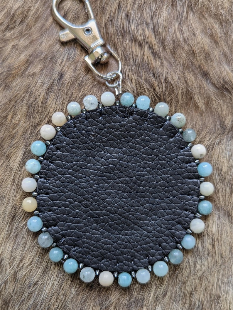 Op de afbeelding: Een ronde zwarte leren sleutelhanger met een zilveren sluiting. De sleutelhanger is versierd met een rand van kleine, ronde, lichtblauwe, witte en gele kralen. De achtergrond is een bruin, getextureerd oppervlak.