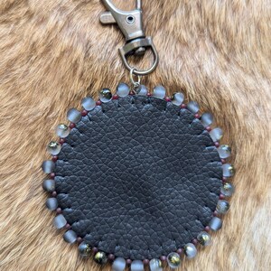 May include: A circular keychain featuring a textured black leather center piece stitched with dark thread. The perimeter is decorated with alternating frosted translucent beads and small dark metallic beads, attached to a silver-toned metal lobster clasp.