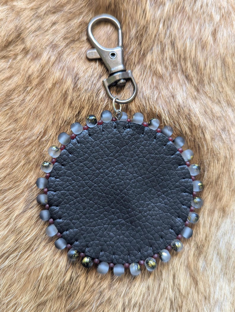 May include: A circular keychain featuring a textured black leather center piece stitched with dark thread. The perimeter is decorated with alternating frosted translucent beads and small dark metallic beads, attached to a silver-toned metal lobster clasp.