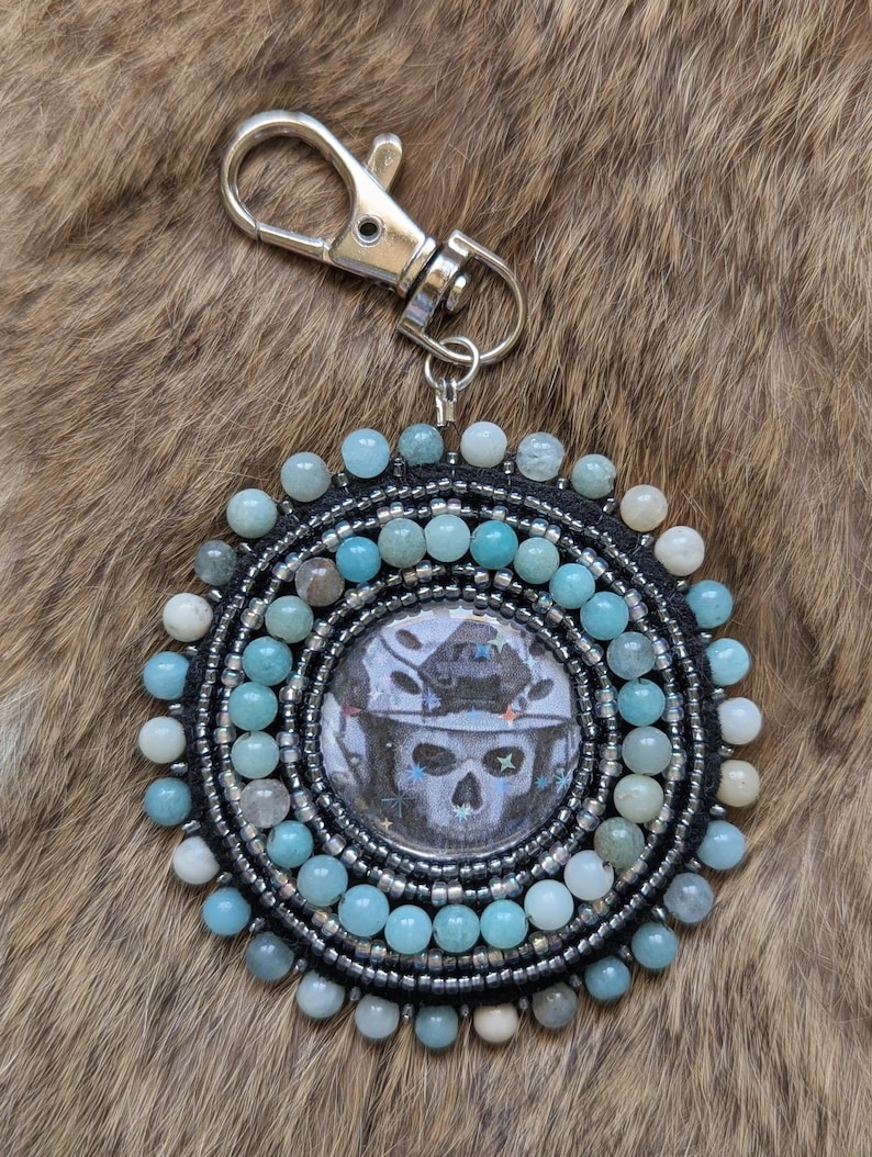 Op de afbeelding: Een ronde, met kralen bezette hanger voor een sleutelhanger, met een schedelontwerp. De hanger heeft lichtblauwe en zilveren kralen en een zwarte kralenrand. Een zilveren sluiting is bevestigd voor gemakkelijk gebruik.