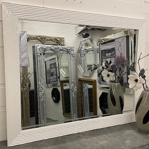 May include: A white, wavy framed mirror with a silver ornate frame reflecting other mirrors in the background. The mirror is sitting on a grey surface with a grey vase and a white flower.