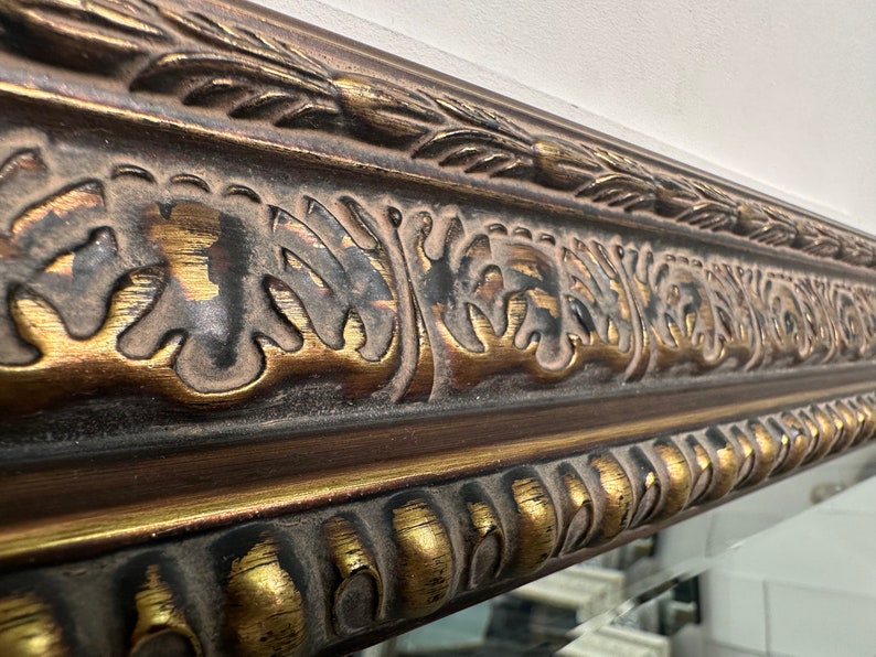 May include: Close-up of a decorative mirror frame with intricate gold and brown detailing. The frame features a raised, ornate design with a textured finish.