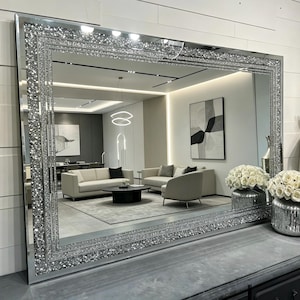 May include: A large rectangular wall mirror featuring a wide, decorative frame filled with crushed silver crystals. The mirror has a smooth, reflective centre surface and a multi-layered border design that catches light, creating a sparkling effect.
