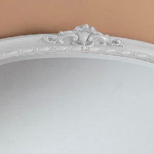 Może przedstawiać: Białe owalne lustro z ozdobną, bogato zdobioną ramą. Rama ma białe, postarzane wykończenie i wypukły wz&oacute;r kwiatowy.