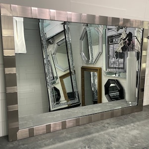 May include: A large, rectangular mirror with a silver-toned frame. The frame has a textured, diamond-like pattern. The mirror is reflecting other mirrors in the background.
