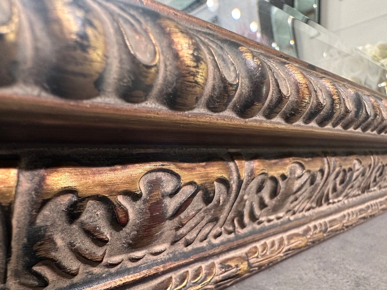 May include: Close-up of a decorative ornate mirror frame with gold and black paint. The frame has a detailed design with intricate carvings and a raised edge.