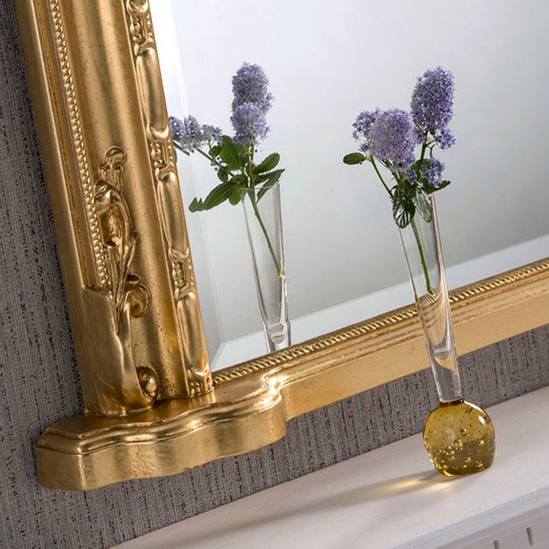 Może przedstawiać: Lustro w złotej ramie z ozdobnymi detalami, z dekoracyjną p&oacute;łką i wazą z fioletowymi kwiatami odbijającymi się w szkle.