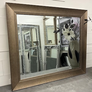 May include: A large, rectangular mirror with a wide, ornate, gold-toned frame. The mirror is reflecting other mirrors and a vase of white flowers.