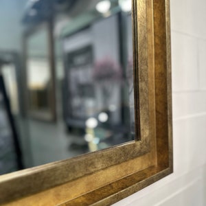 May include: A rectangular wall mirror featuring a wide, stepped frame with a distressed, antique gold finish. The frame has a multi-layered profile that creates depth, and the reflective glass surface shows a blurred interior room reflection.