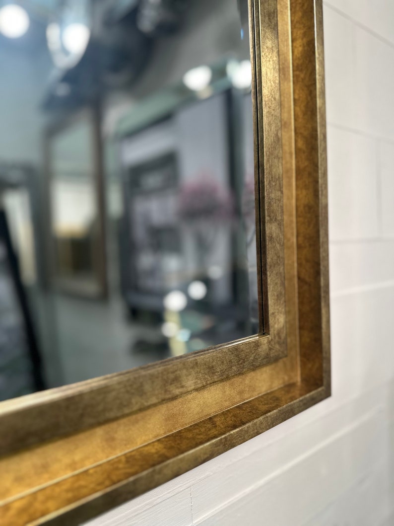 May include: A rectangular wall mirror featuring a wide, stepped frame with a distressed, antique gold finish. The frame has a multi-layered profile that creates depth, and the reflective glass surface shows a blurred interior room reflection.