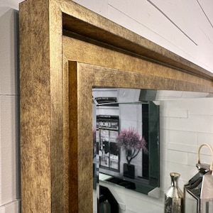 May include: A rectangular wall mirror featuring a wide, textured gold-toned frame with a stepped, multi-layered profile. The glass has a beveled edge, reflecting a room interior with a decorative lantern, a vase, and a floral arrangement.