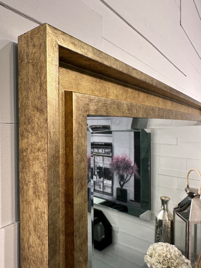 May include: A rectangular wall mirror featuring a wide, textured gold-toned frame with a stepped, multi-layered profile. The glass has a beveled edge, reflecting a room interior with a decorative lantern, a vase, and a floral arrangement.