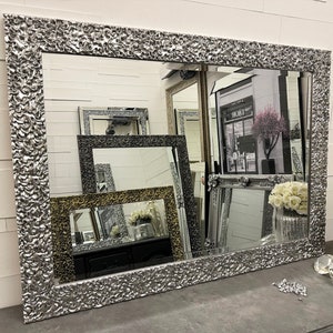 May include: A large silver mirror with a textured frame. The frame has a raised, floral design. The mirror is reflecting other mirrors and a shop interior.