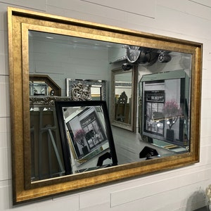 May include: A large rectangular wall mirror featuring a wide, textured gold-toned frame. The reflective surface displays multiple other mirrors and framed art pieces, including a black-framed print with the text DIOR visible in the reflection.