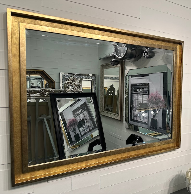 May include: A large rectangular wall mirror featuring a wide, textured gold-toned frame. The reflective surface displays multiple other mirrors and framed art pieces, including a black-framed print with the text DIOR visible in the reflection.
