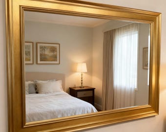 Op de afbeelding: Een grote rechthoekige spiegel met een goudkleurige lijst. De lijst heeft een gelaagd ontwerp. De spiegel reflecteert een slaapkamer met een bed, een nachtkastje, een lamp en een raam met gordijnen. De spiegel is aan een witte muur bevestigd.