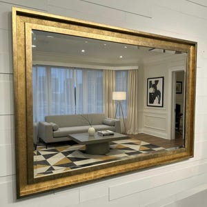 May include: A large rectangular wall mirror featuring a wide, textured gold frame with a distressed finish. The mirror reflects a living room interior containing a light grey sofa, a square coffee table, a floor lamp, and a geometric rug.