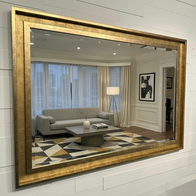 May include: A large rectangular wall mirror featuring a wide, textured gold frame with a distressed finish. The mirror reflects a living room interior containing a light gray sofa, a square coffee table, a floor lamp, and a geometric rug.