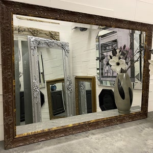 May include: A large, ornate, brown wood framed mirror with a gold-toned inner frame. The mirror reflects other mirrors and a vase of white flowers.