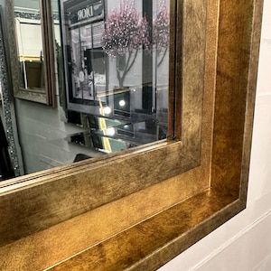May include: A rectangular wall mirror featuring a wide, textured frame with a distressed antique gold finish. The frame has a stepped, multi-layered profile that creates depth. The reflective glass surface shows a partial reflection of a storefront.