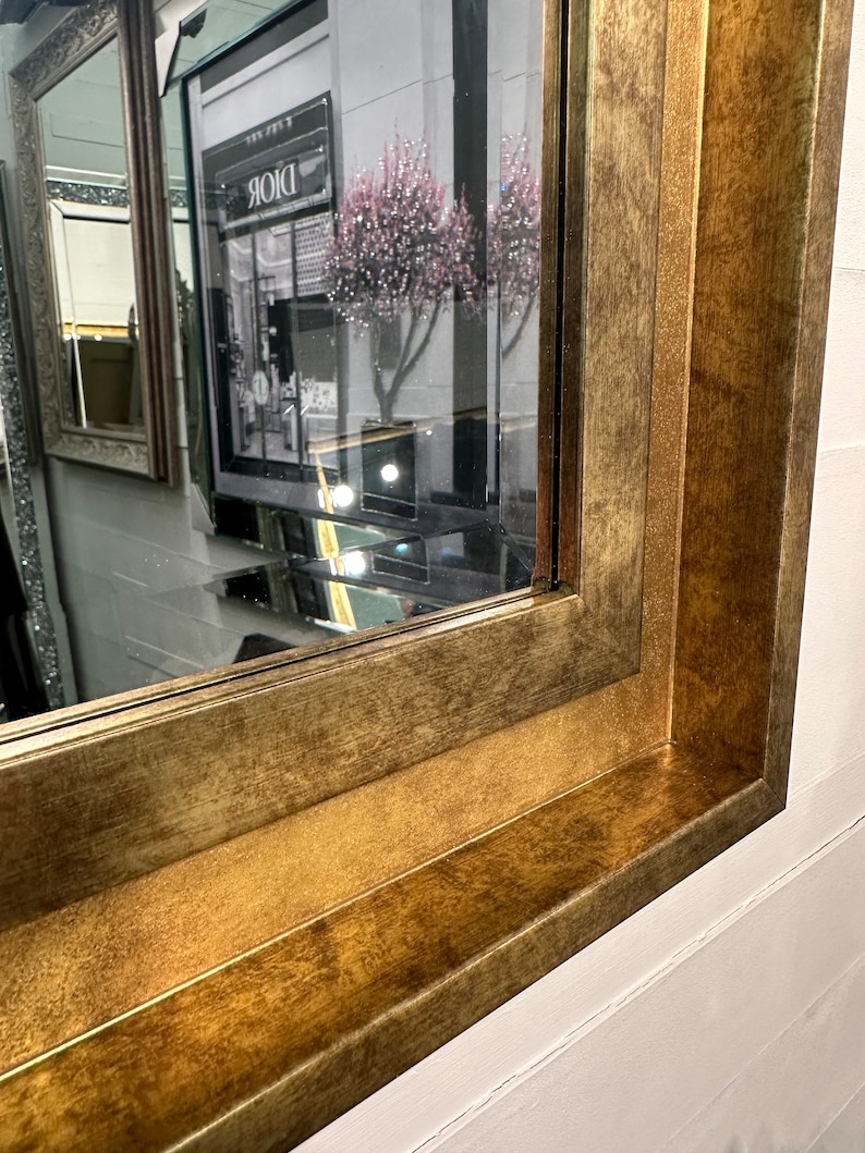 May include: A rectangular wall mirror featuring a wide, textured frame with a distressed antique gold finish. The frame has a stepped, multi-layered profile that creates depth. The reflective glass surface shows a partial reflection of a storefront.