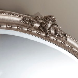 Może przedstawiać: Srebrne owalne lustro z ozdobną, szczeg&oacute;łową ramą. Rama ma dekoracyjny wz&oacute;r z motywem zwijania.