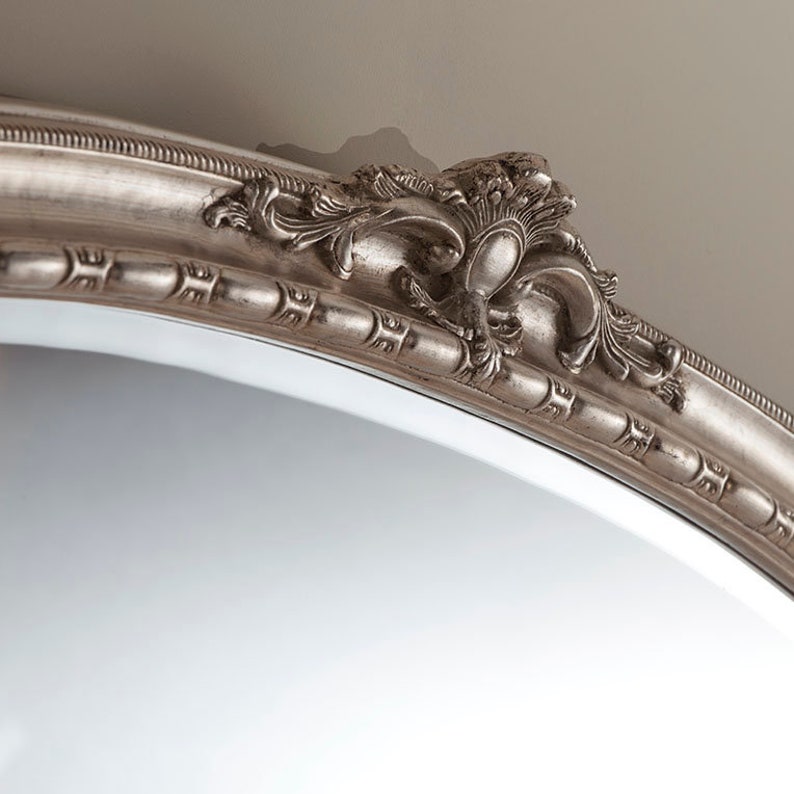 Może przedstawiać: Srebrne owalne lustro z ozdobną, szczeg&oacute;łową ramą. Rama ma dekoracyjny wz&oacute;r z motywem zwijania.