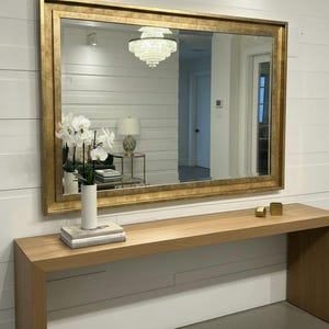 May include: A large rectangular wall mirror featuring a wide, textured gold frame. The mirror is mounted horizontally on a white shiplap wall above a long, light-toned wooden console table that holds a white vase with white orchid flowers.