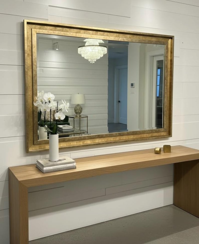 May include: A large rectangular wall mirror featuring a wide, textured gold frame. The mirror is mounted horizontally on a white shiplap wall above a long, light-toned wooden console table that holds a white vase with white orchid flowers.